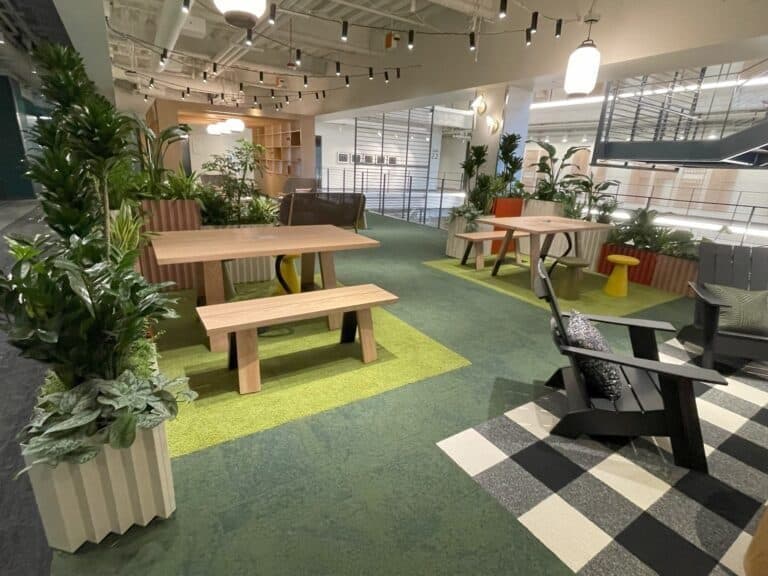 Office commons with a long communal picnic table framed by biophilic planting.