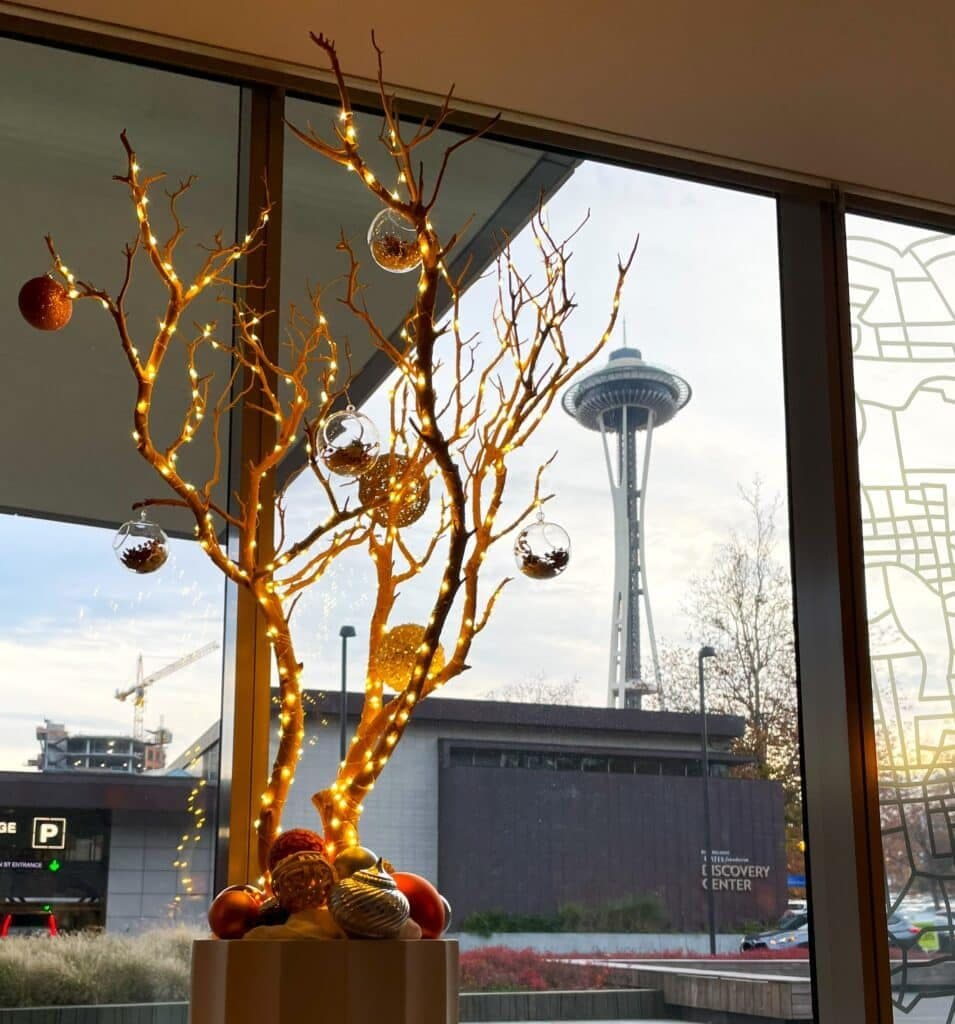 Illuminated golden branch display hung with ornaments beside a window overlooking the Seattle Space Needle at dusk.
